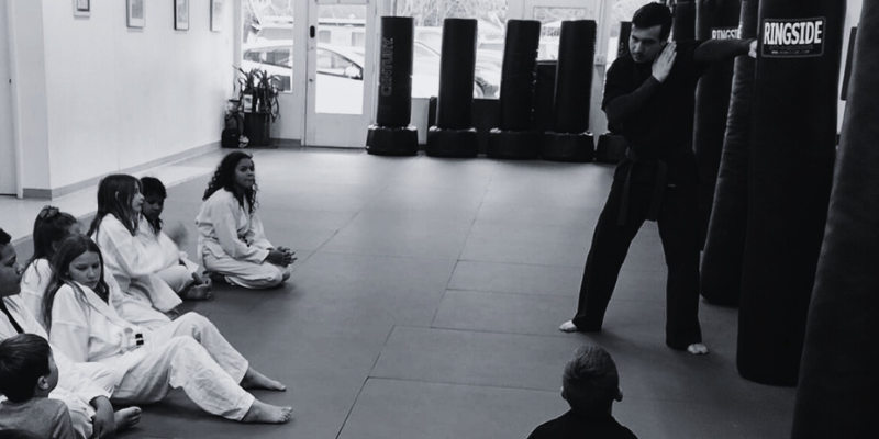 Elevate students seated on the mat watching an instructor demonstrate a technique.